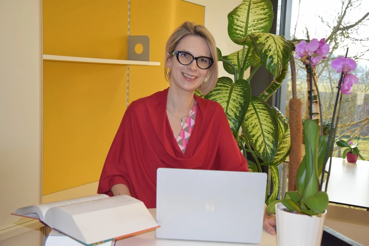 FokusDEMENZ - Radka Eder sitzt auf einem Stuhl mit einem Laptop und einem Buch auf dem Schreibtisch